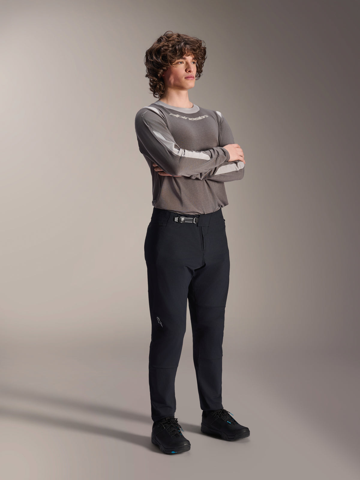 Mannelijk model draagt Alpinestars A-Dura Elite broek, zwarte MTB-broek, gecombineerd met een grijze jersey met lange mouwen en zwarte fietsschoenen, ratelsluitsysteem aan de voorzijde, professionele mountainbikekleding voor trail- en enduroritten