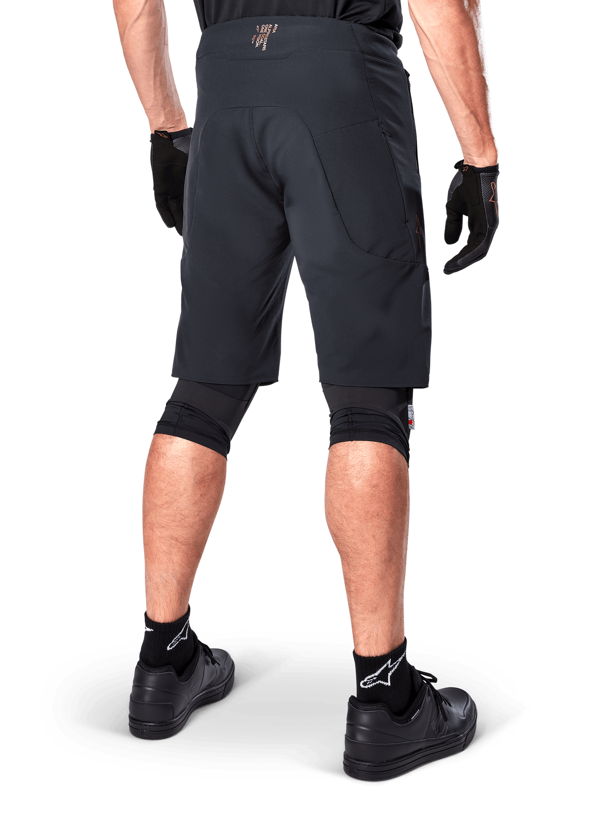 Alpinestars A-Aria Elite Shorts in het zwart, prestatiegerichte mountainbike short, achteraanzicht van een fietser met zwarte fietshandschoenen en zwarte schoenen, ontworpen voor optimale bewegingsvrijheid op off-road trails