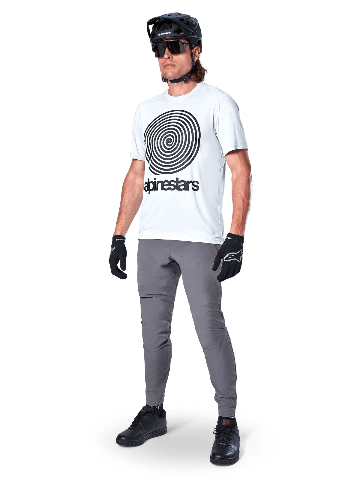 Alpinestars A-Dura broek, MTB-broek, donkergrijs, gedragen met wit T-shirt met spiraalprint, zwarte MTB-helm, zonnebril en zwarte handschoenen, duurzame 2-way stretch stof voor off-road fietsen