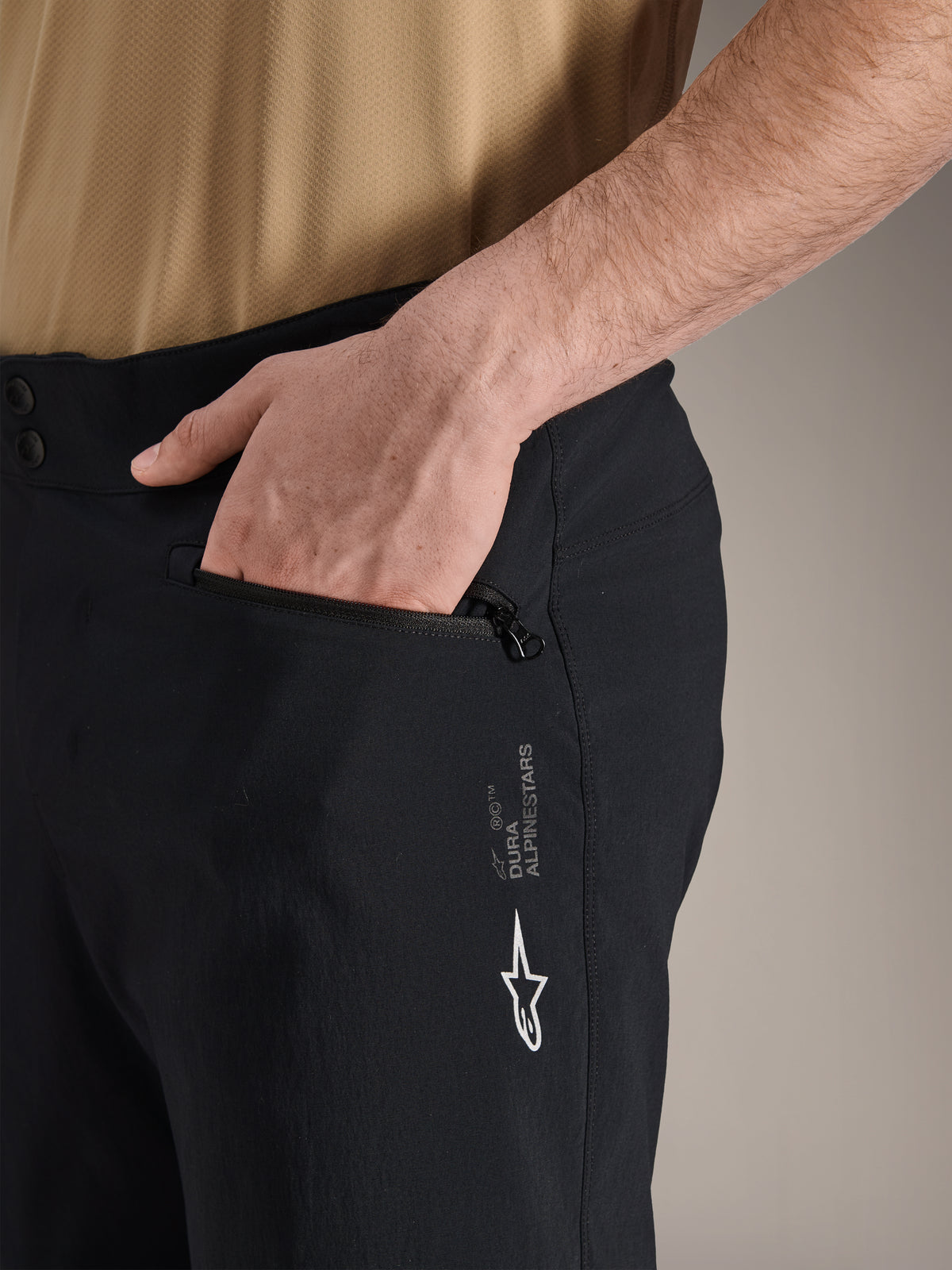Alpinestars A-Dura Shorts, zwarte mountainbikeshorts, close-up van zijzak met ritssluiting en hand erin, voorzien van wit Alpinestars-logo en A-Dura-branding, duurzaam textiel voor off-road fietsen