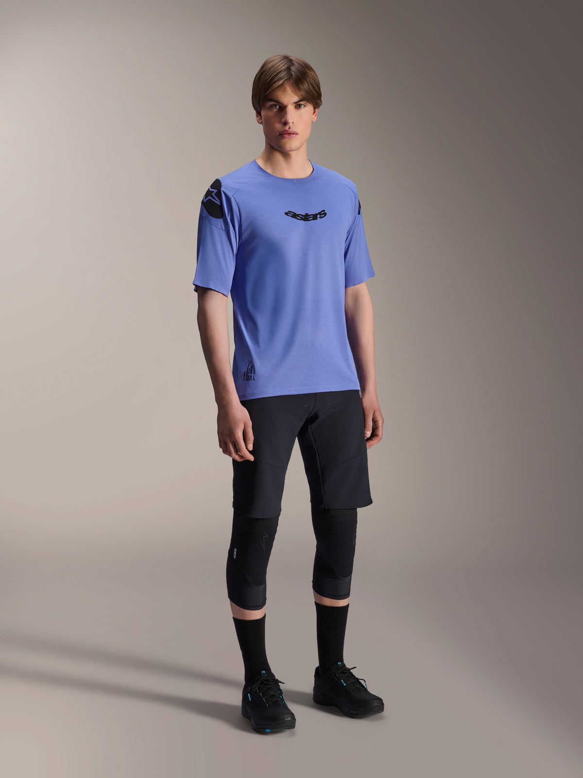 Mannelijk model draagt Alpinestars A-Aria Polartec Elite Short Sleeve Jersey in Baja Blue, gecombineerd met een zwarte MTB-short en kniebeschermers, voorzien van zwarte Astars-logo's op de borst en schouder, hoogwaardige mountainbike-uitrusting