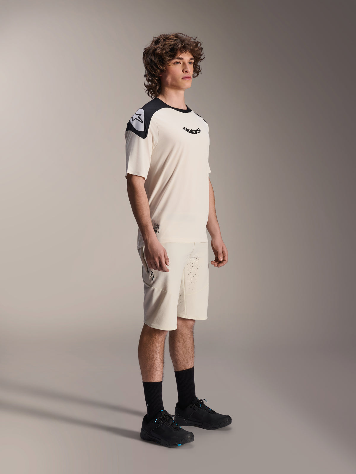 Mannelijk model in Alpinestars A-Aria Elite Shorts in Ecru, technische mountainbikeshorts met lasergesneden ventilatie, gecombineerd met een bijpassend crème en zwart fietsshirt met korte mouwen en zwarte schoenen, professionele wielerkleding.