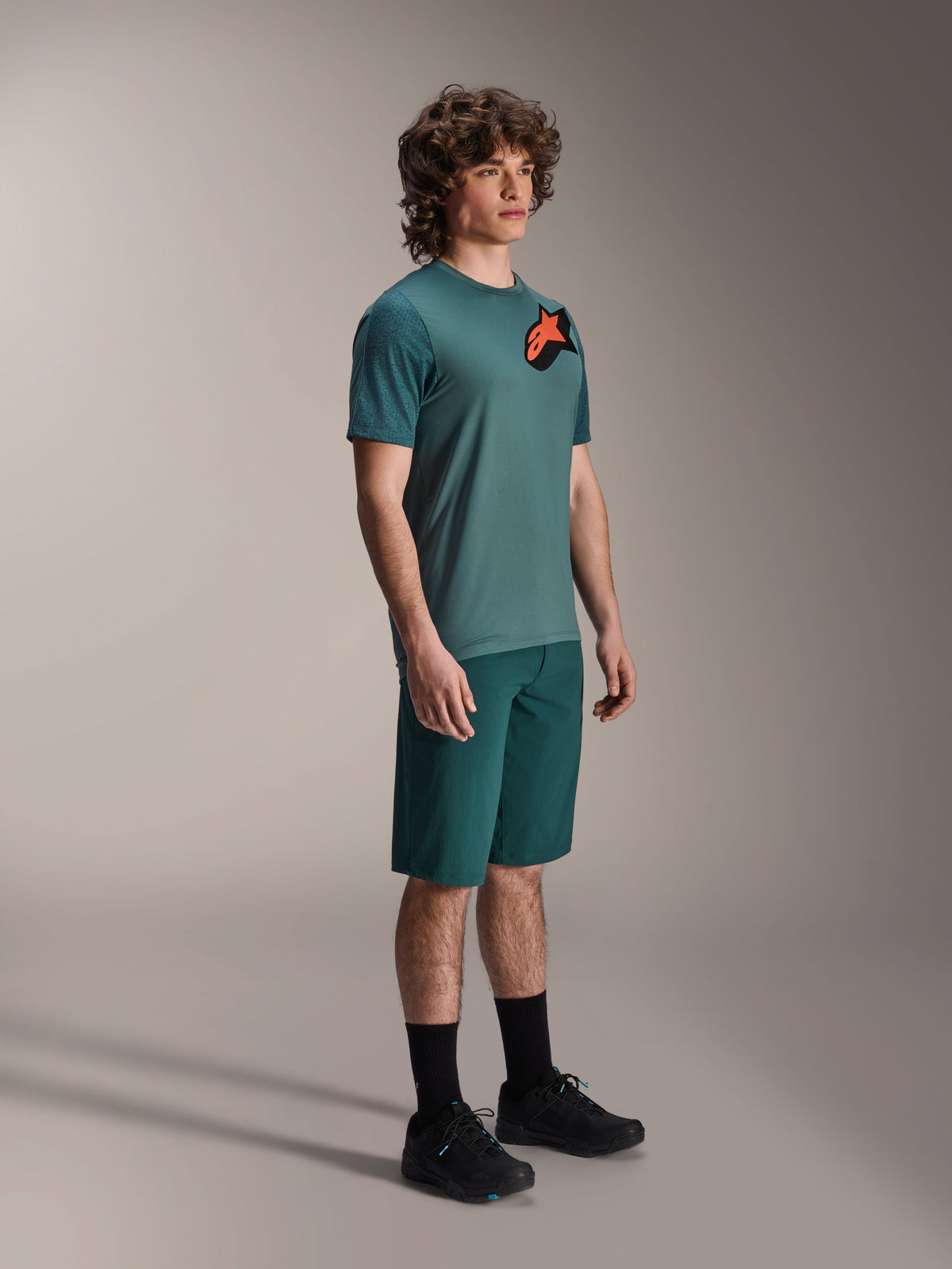 Alpinestars A-Dura Lipan Jersey, MTB-shirt met korte mouwen, Silver Pine groen, met een oranje en zwart Astars-logo op de borst, gedragen met bijpassende groene shorts en zwarte fietsschoenen, casual trailstijl