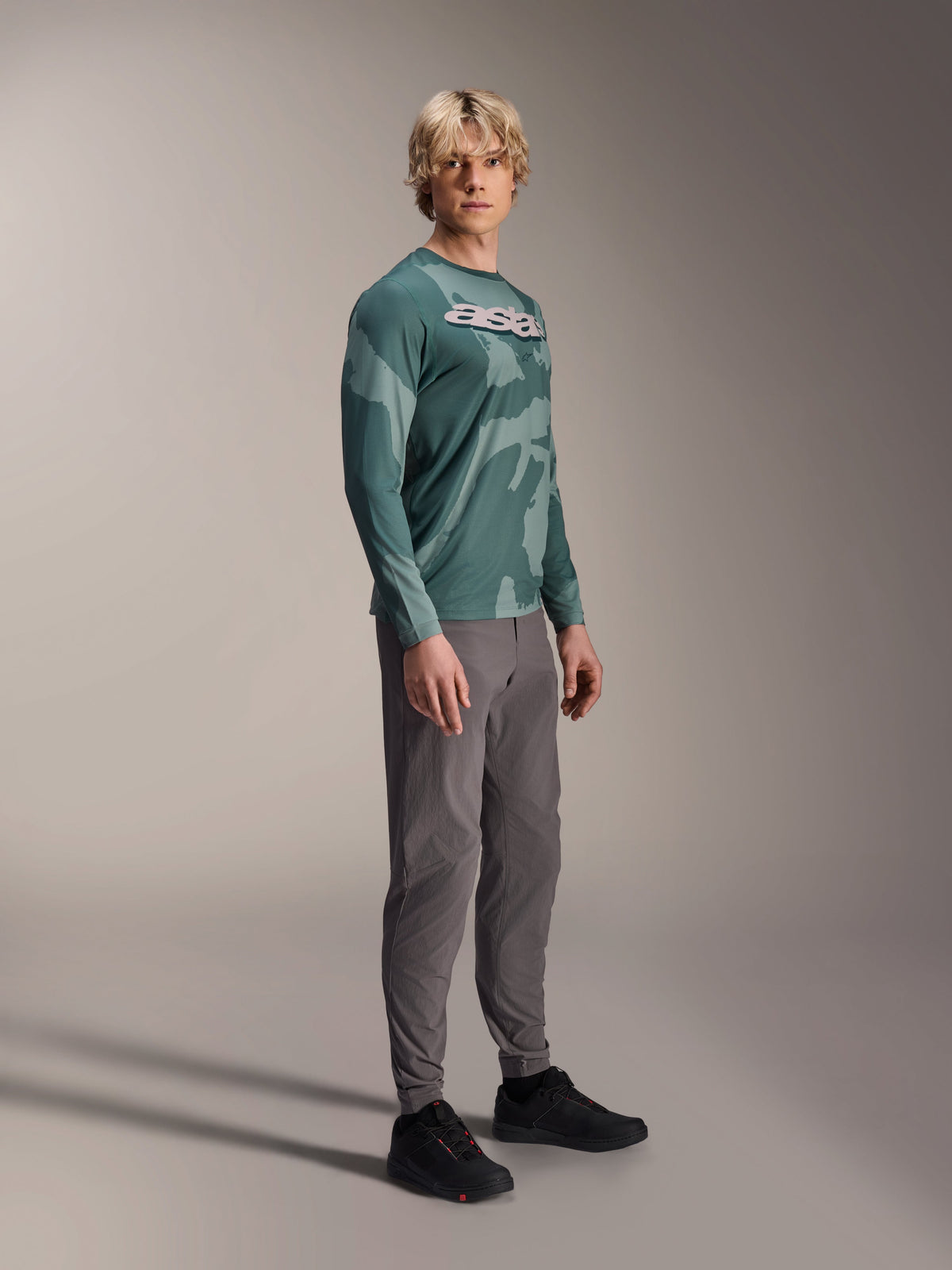 Alpinestars A-Dura Hollow Camo Jersey met lange mouwen, Silver Pine Camo, gedragen door een model met een grijze broek en zwarte schoenen, voorzien van een groen camouflagepatroon en een opvallend wit ASTARS-logo op de borst, hoogwaardige MTB-kleding