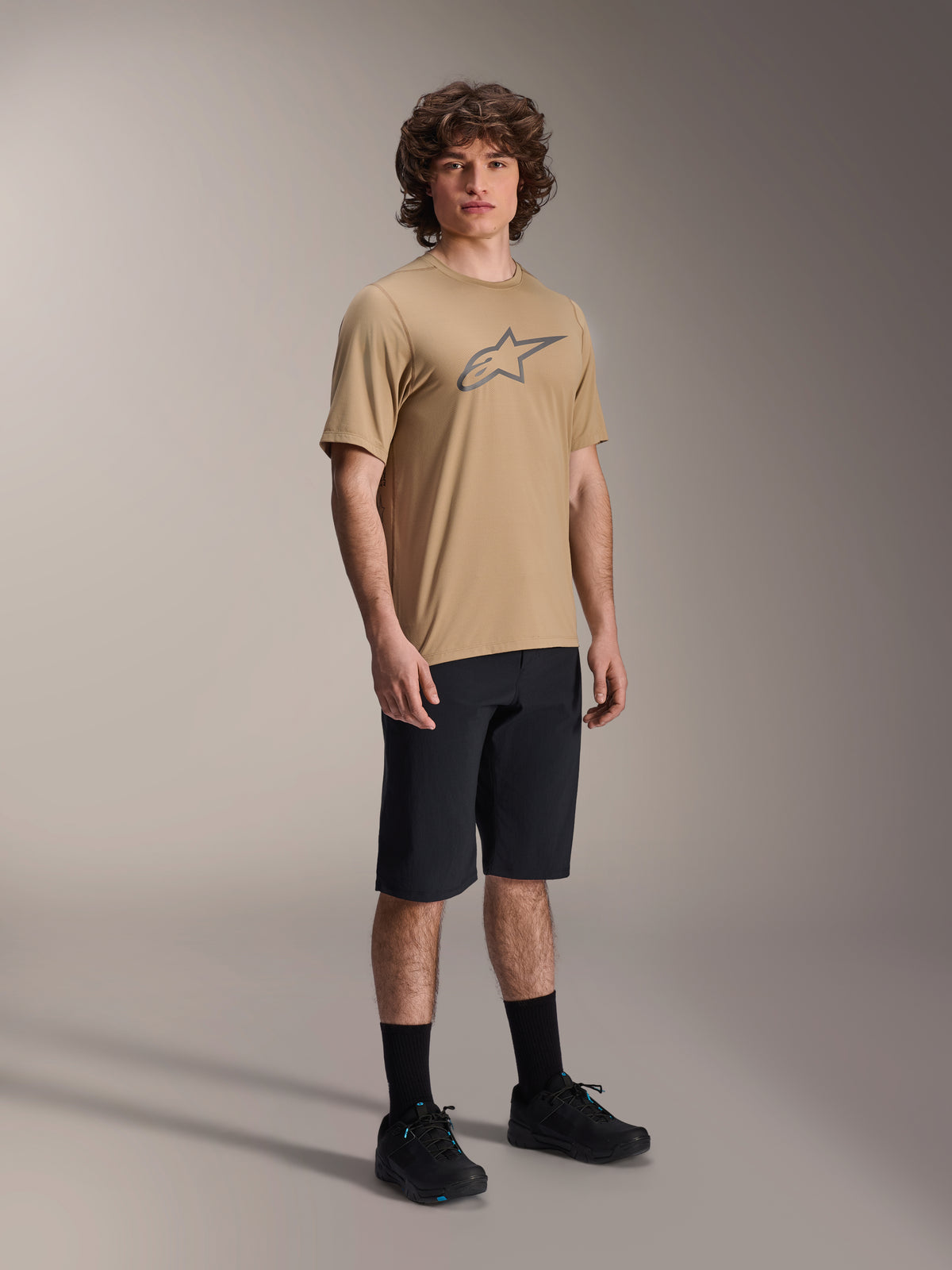 Alpinestars A-Dura Shorts, zwarte mountainbike short, gedragen door een model met een beige jersey met korte mouwen en het Alpinestars-logo, zwarte sokken en zwarte schoenen, duurzame MTB-uitrusting voor off-road ritten