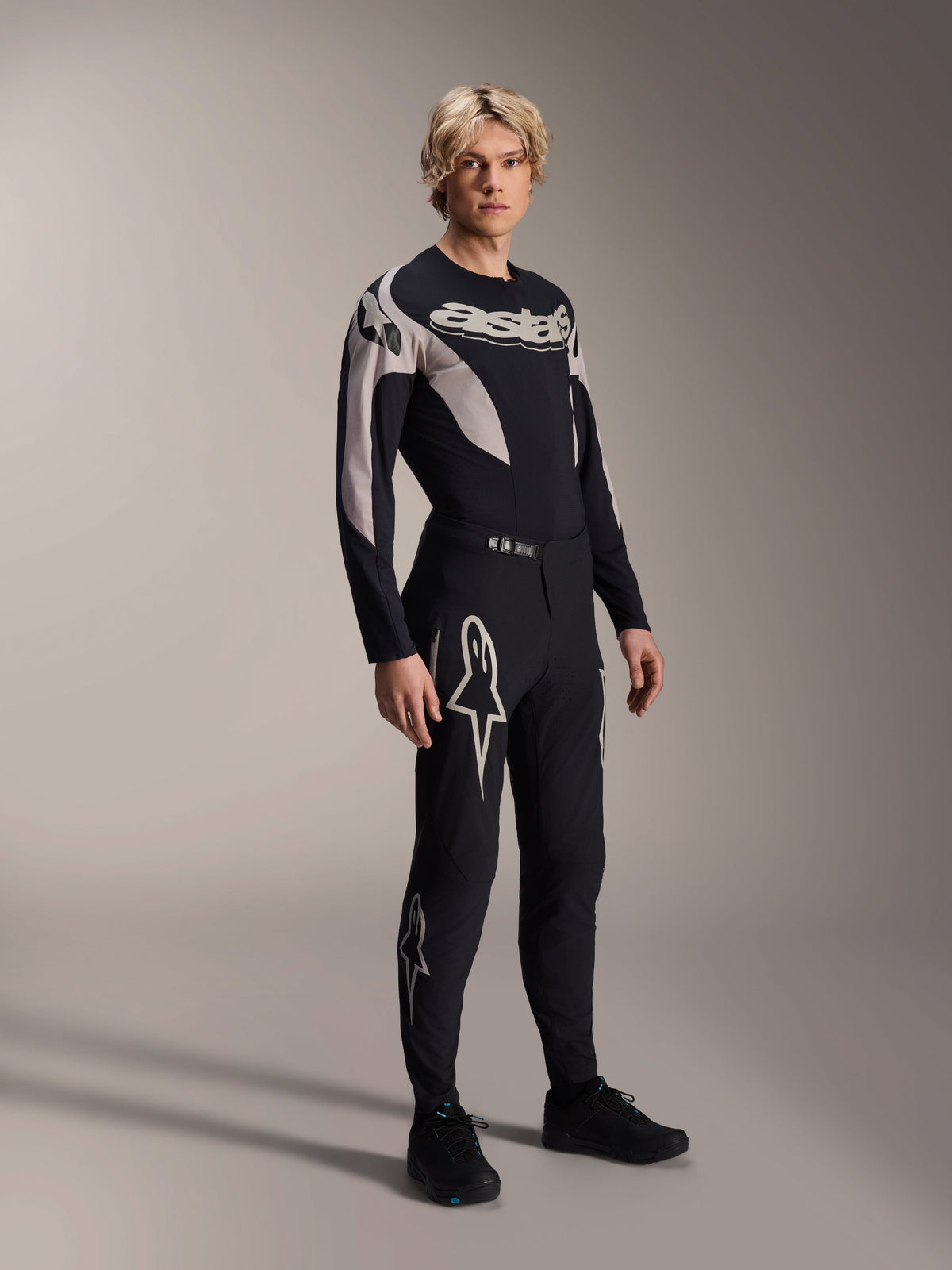 Mannelijk model draagt Alpinestars A-Supra Melt Jersey met lange mouwen, zwart met grijze grafische accenten, 'astars' logo op de borst, gecombineerd met bijpassende zwarte broek en schoenen, performance mountainbike-uitrusting voor off-road racen