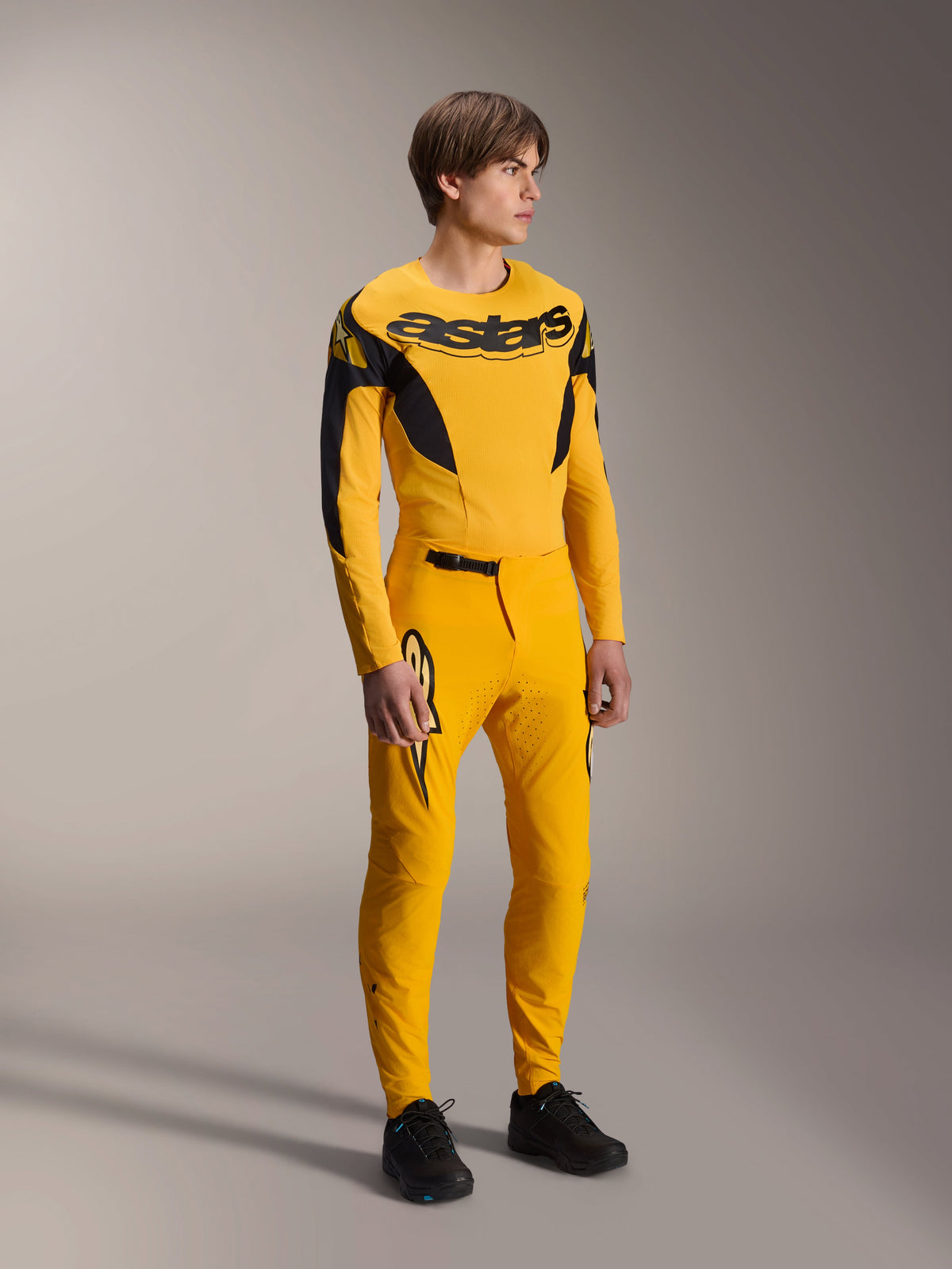 Model draagt Alpinestars Limited Edition A-Supra Melt Gold Jersey, MTB-jersey met lange mouwen in donkergeel en zwart, gecombineerd met bijpassende gele broek en zwarte schoenen, technische off-road fietskleding met racing-geïnspireerde graphics