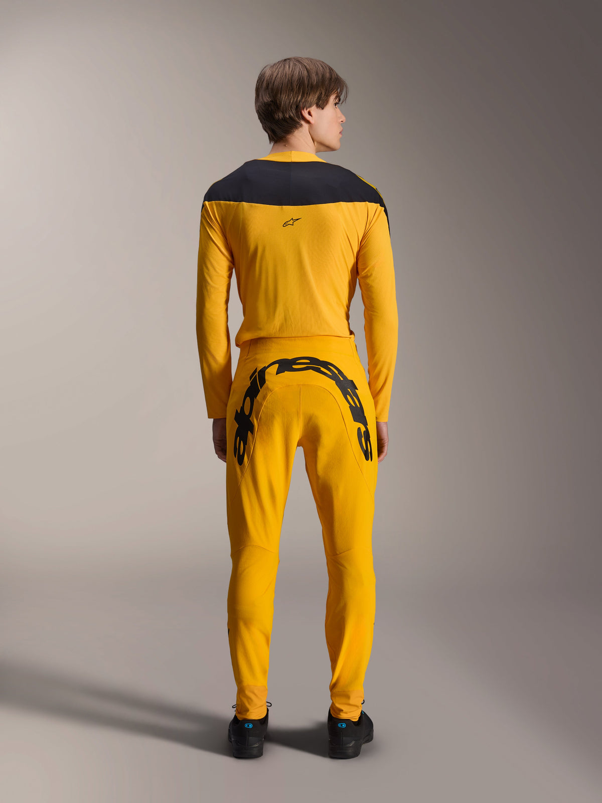 Alpinestars Limited Edition A-Supra Melt Gold Jersey, MTB-shirt met lange mouwen, donkergeel, achteraanzicht, met zwarte schouderpanelen, gedragen met bijpassende gele broek met grote zwarte merknaam op de achterzijde, performance fietskleding