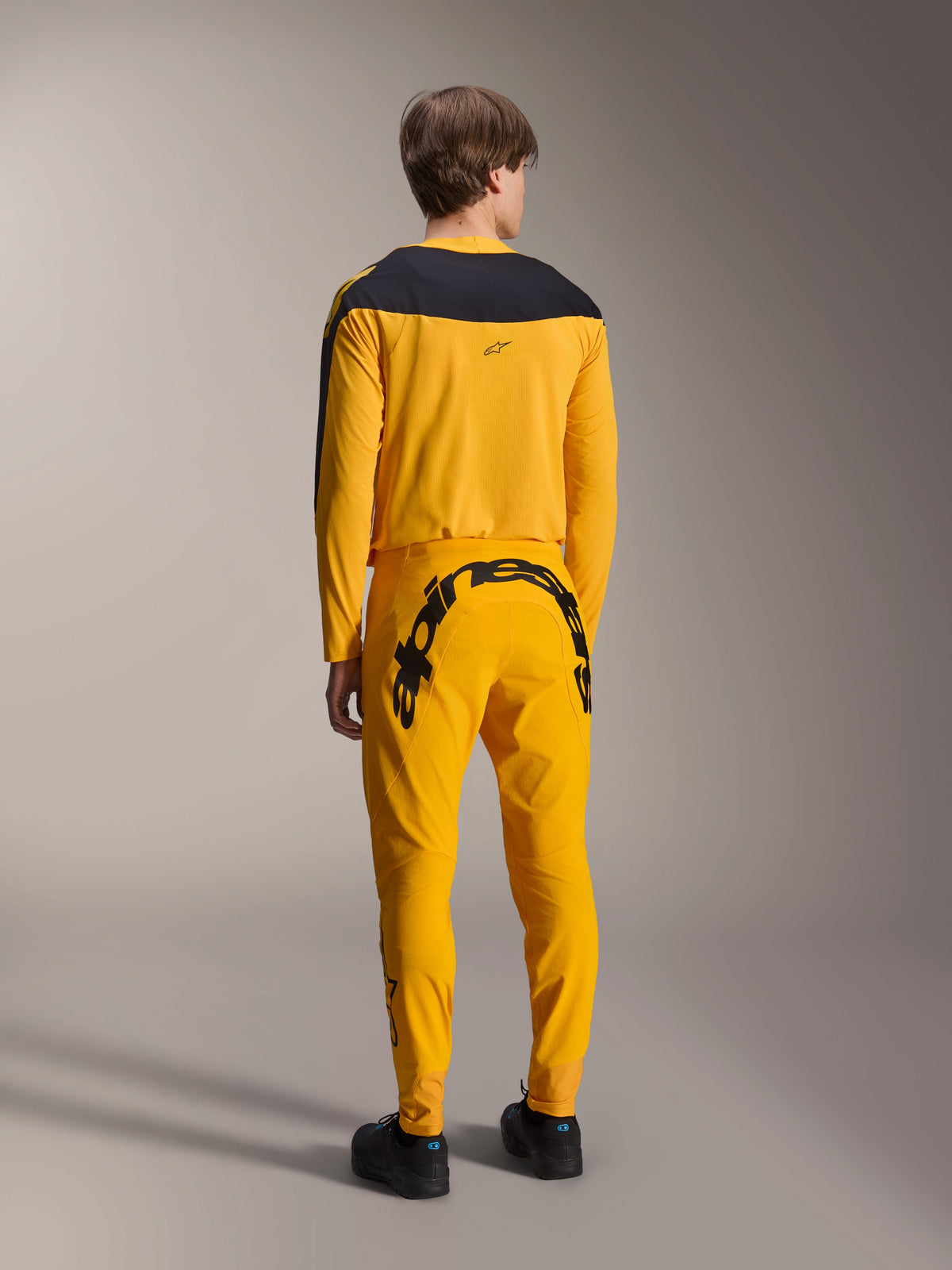 Alpinestars Limited Edition A-Supra Melt Gold Jersey, MTB-jersey met lange mouwen, donkergeel met zwart schouderpaneel, gedragen met bijpassende gele broek met opvallende zwarte branding op de achterzijde, achteraanzicht van prestatiegerichte fietsuitrusting