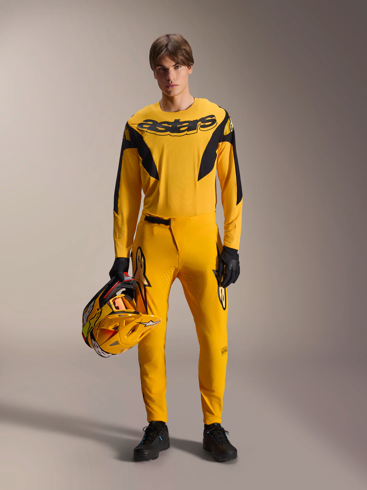 Model draagt Alpinestars Limited Edition A-Supra Melt Gold MTB-kit, gele jersey met lange mouwen en zwart 'astars'-logo, bijpassende gele broek met zwarte accenten, houdt een geel-zwarte racehelm vast, zwarte handschoenen en schoenen, performance-uitrusting voor mountainbiken