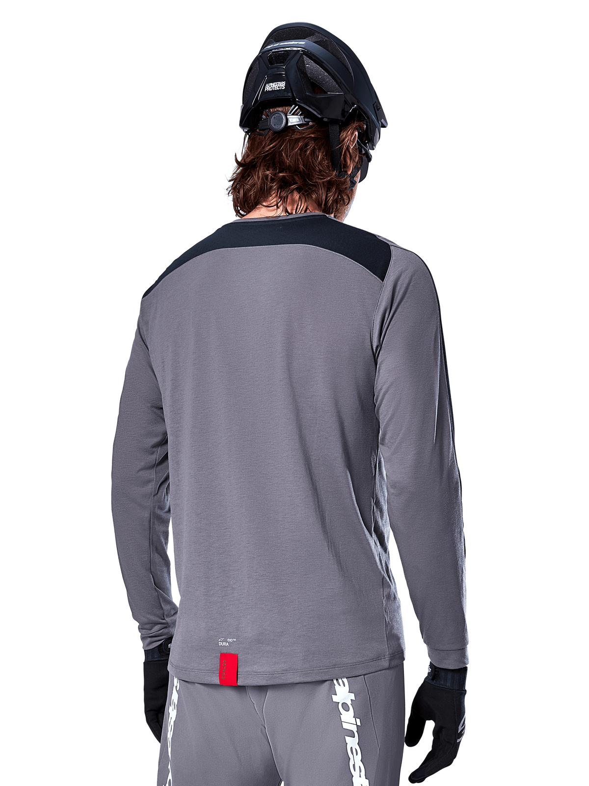 Alpinestars A-Dura Dri Astar Long Sleeve Jersey, Dark Gray MTB jersey shown from the back, featuring a black shoulder panel and small red brand tag, worn with a black mountain bike helmet and gloves.
