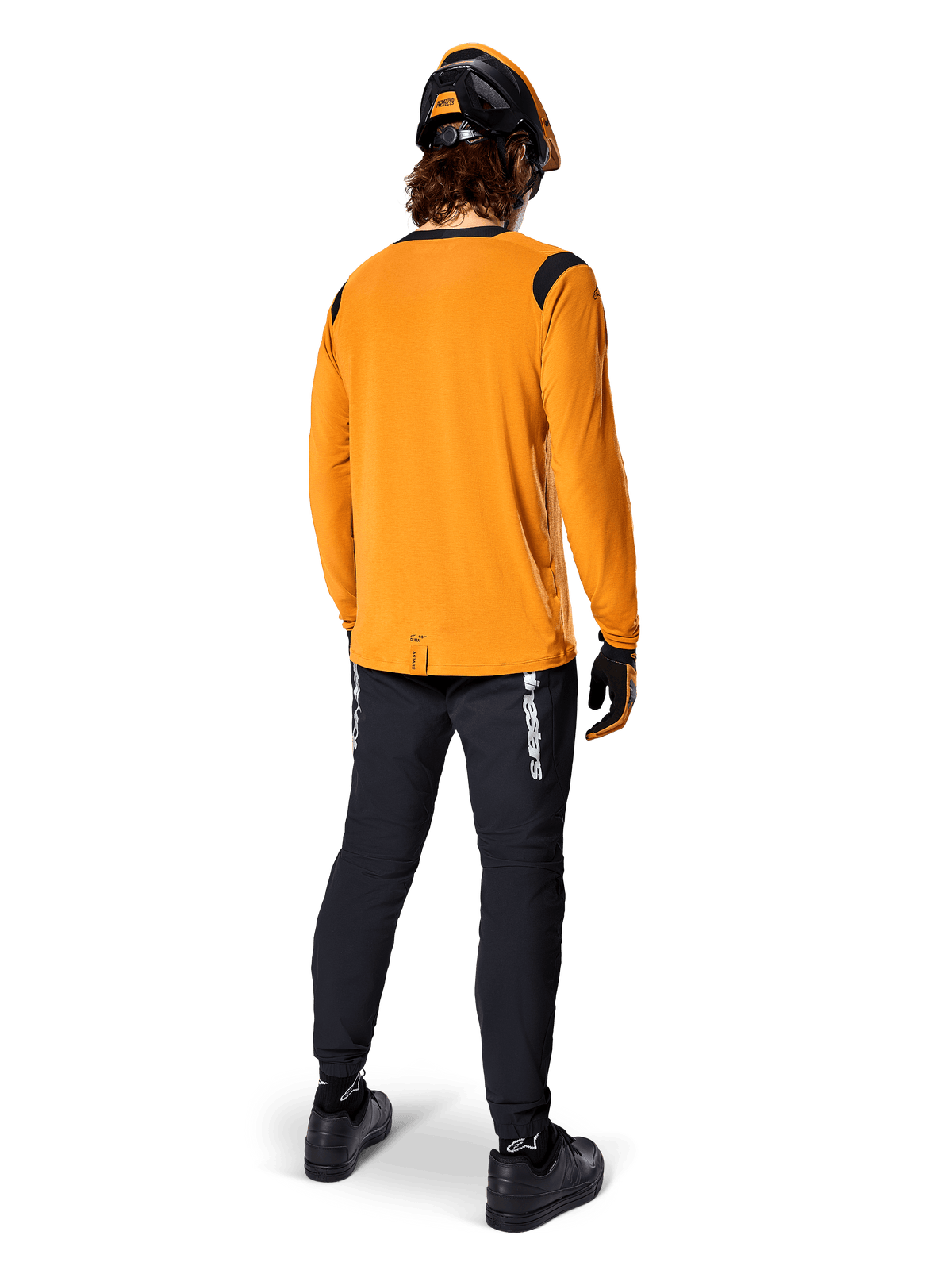 Mountainbiker van achteren gezien, dragend het Alpinestars A-Dura Dri Wool Jersey met lange mouwen in Dark Gold, zwarte technische broek met witte logo's en een bijpassende zwart-oranje helm, prestatiegerichte mountainbikekleding.