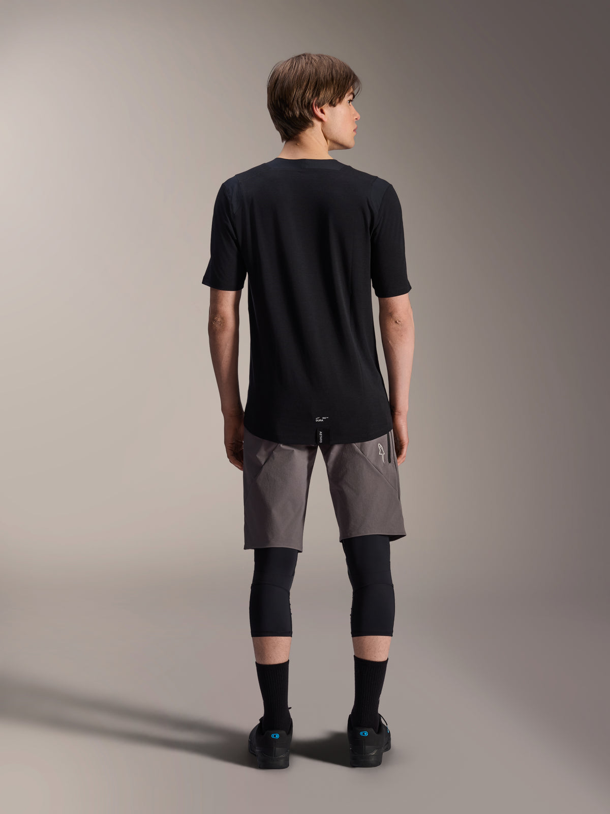 Alpinestars A-Dura Dri Wool Jersey met korte mouwen, zwart, achteraanzicht gedragen door model met grijze MTB-short en zwarte kniebeschermers, voorzien van een langere achterzijde en atletische pasvorm voor off-road fietsen