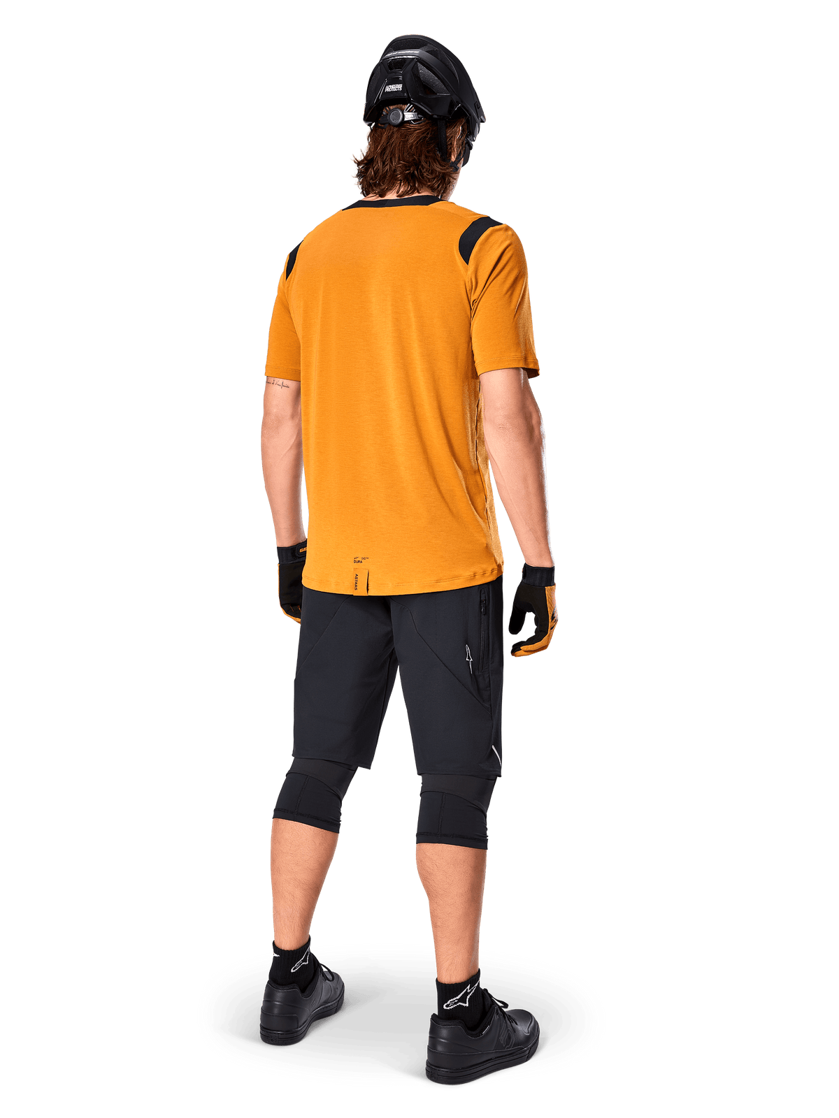 Alpinestars A-Dura Dri Wool Jersey met korte mouwen, fietsshirt, Dark Gold, achteraanzicht van mountainbiker in mosterdgeel shirt met zwarte mesh schouderaccenten, gecombineerd met zwarte short, kniebeschermers, zwarte helm en handschoenen, technische off-road wieleruitrusting