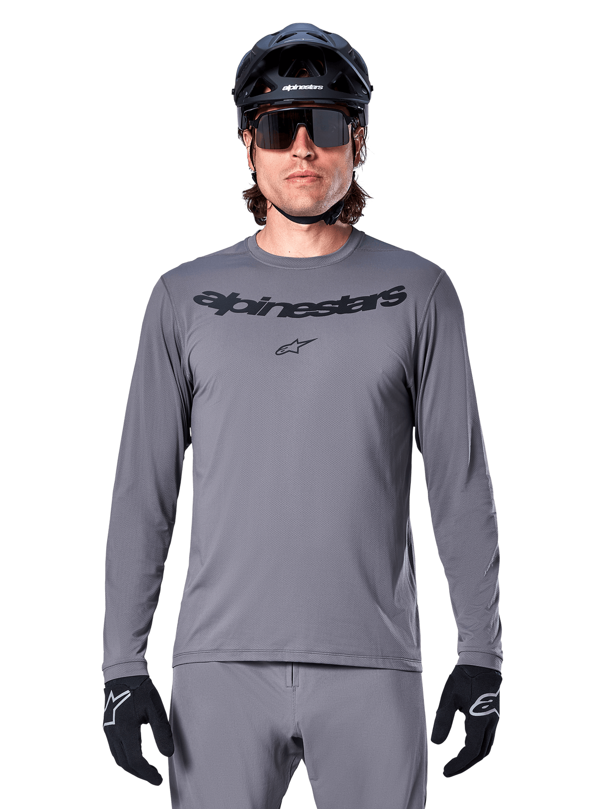 Mannelijke rijder in het Alpinestars A-Dura Rocker Jersey met lange mouwen, donkergrijze MTB-jersey met zwart woordmerk op de borst, gecombineerd met grijze broek, zwarte helm en handschoenen, ontworpen voor off-road fietsen