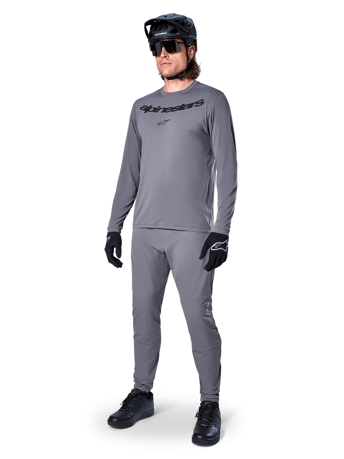 Alpinestars A-Dura Rocker Jersey met lange mouwen, donkergrijs, gedragen door een model met bijpassende grijze broek, zwarte mountainbikehelm, zonnebril en handschoenen, casual MTB-stijl voor off-road rijden