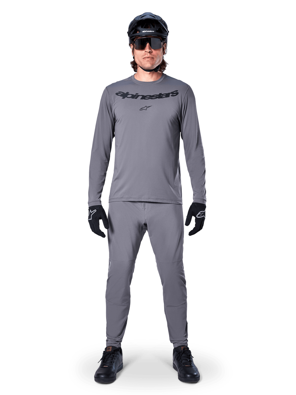 Mannelijke fietser in Alpinestars A-Dura Rocker Jersey met lange mouwen in donkergrijs, bijpassende grijze broek, zwarte MTB-helm, zonnebril en handschoenen, professionele mountainbikekleding voor off-road paden