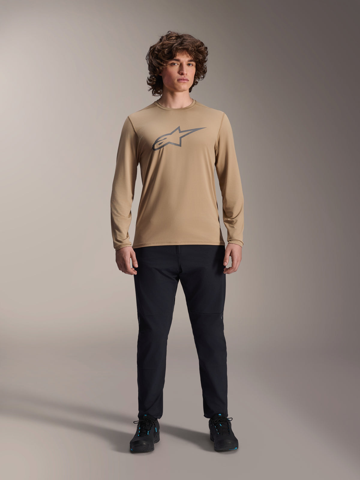 Alpinestars A-Dura Astar Jersey met lange mouwen, lichtbruin met grijs Astars-logo op de borst, gedragen door een model met een zwarte technische broek en schoenen, casual mountainbike-prestatiekleding