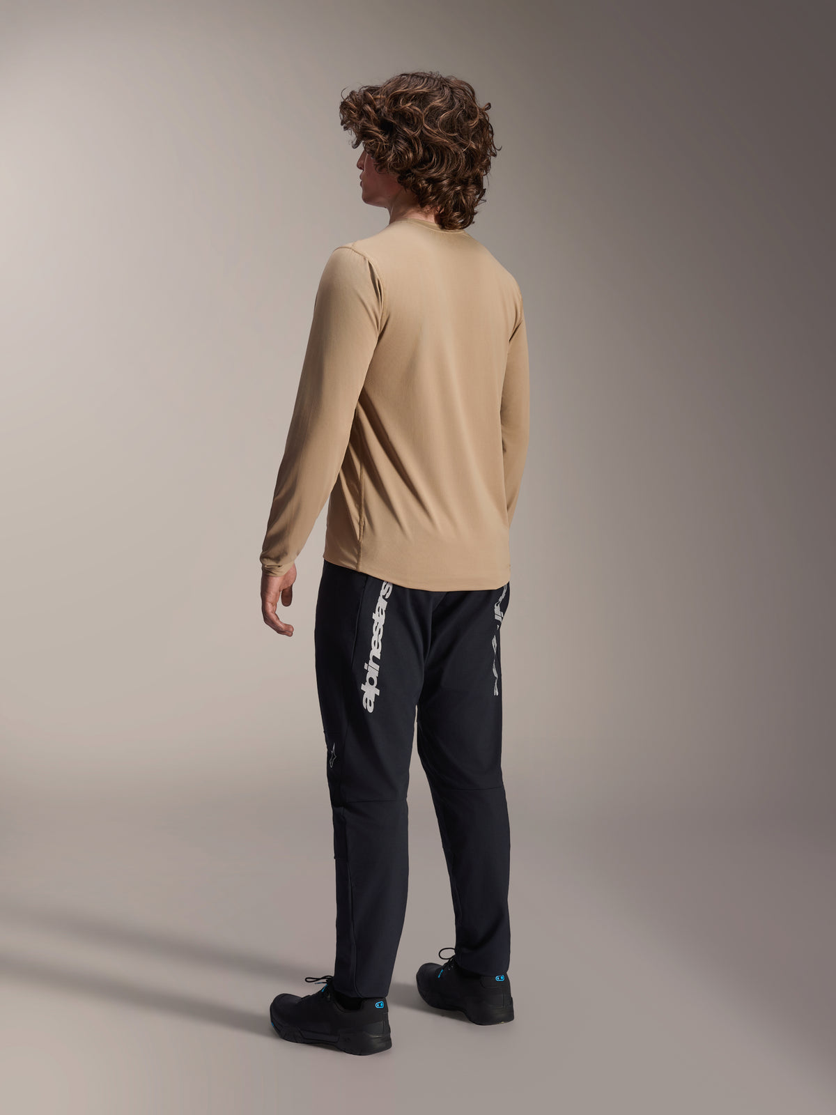 Alpinestars A-Dura Astar Long Sleeve Jersey in lichtbruin, achteraanzicht van een mannelijk model toont het drop tail-ontwerp, gecombineerd met zwarte broek en schoenen, casual mountainbike-prestatiekleding