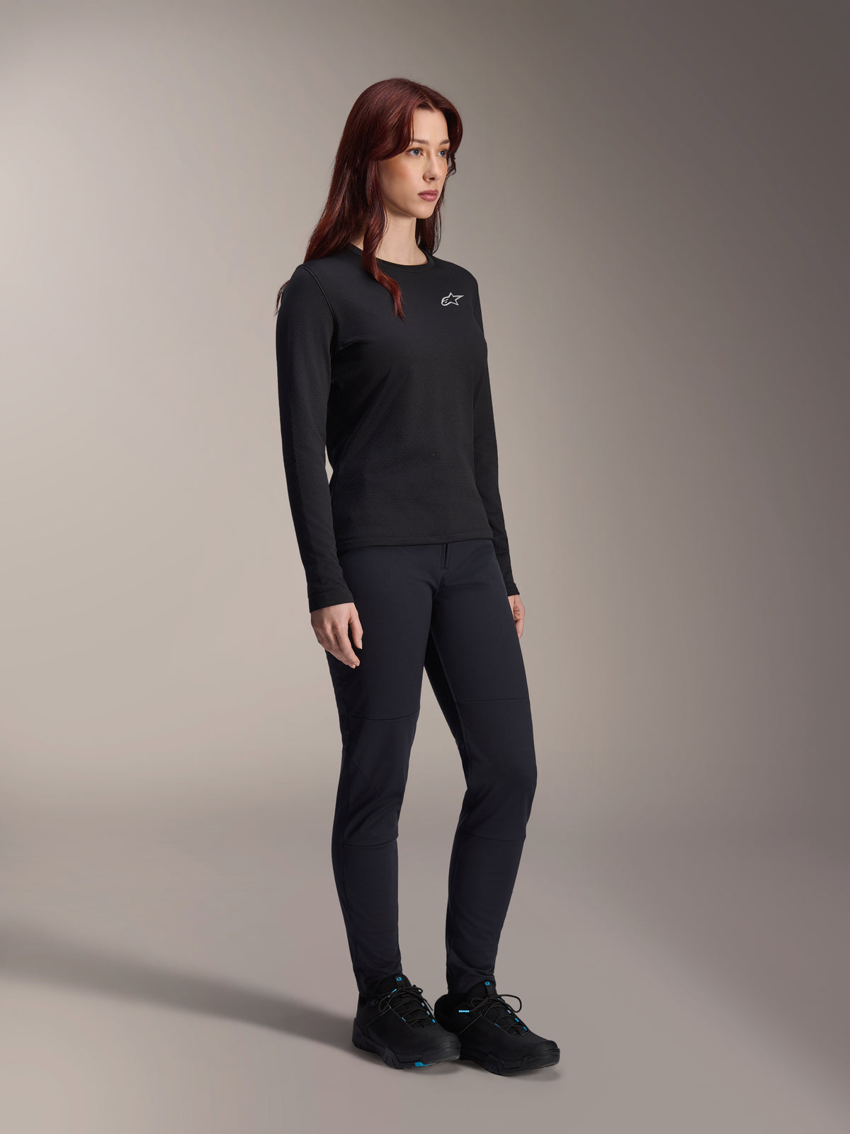 Vrouw die de Alpinestars Stella A-Dura Thermal Jersey draagt, zwart MTB-shirt met lange mouwen en wit logo op de borst, gestyled met bijpassende zwarte broek en sportschoenen, studioportret voor technische wielerkleding