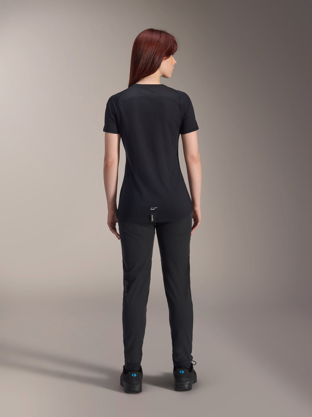 Alpinestars Stella A-Dura Dri Switch Jersey voor dames met korte mouwen, zwart, achteraanzicht op een model, voorzien van een verlengde achterkant en technische mesh-panelen, gecombineerd met een zwarte broek, performance MTB-wielerkleding