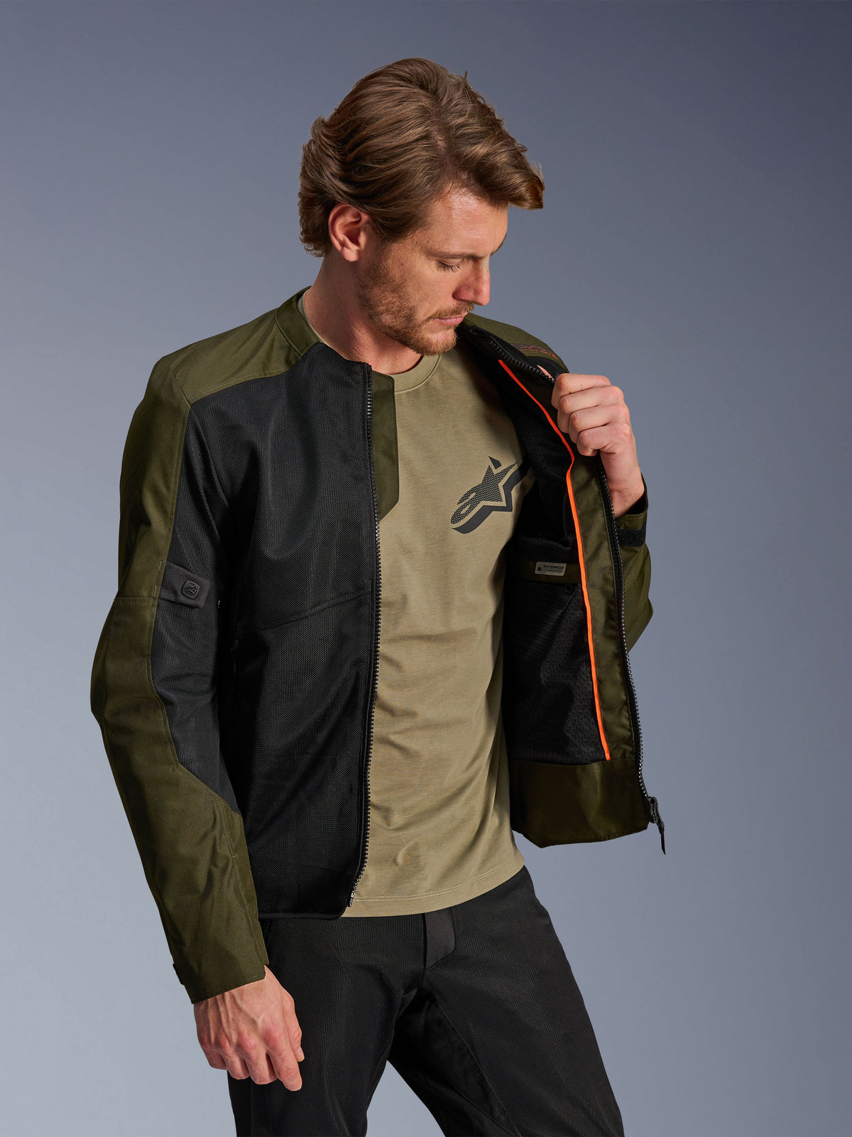 Alpinestars C-1 Air Jacket in Khaki en zwart, gedragen door een mannelijk model om de oranje binnenvoering en het waterdichte compartiment te tonen, voorzien van grote tech mesh-panelen voor luchtstroom, urban eigentijdse motorstijl gecombineerd met een bijpassend olijfgroen t-shirt en zwarte broek