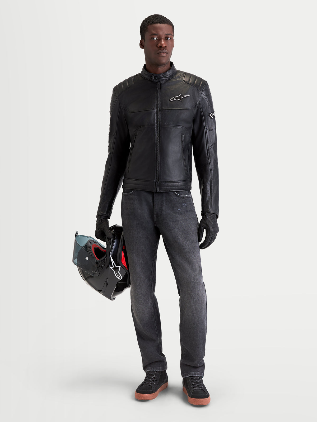 Alpinestars Revern leren jas, heren motorjas voor urban rijden in zwart, vooraanzicht van een model met donkere jeans en een helm in de hand, met verweerd rundleer en heritage bikerstijl