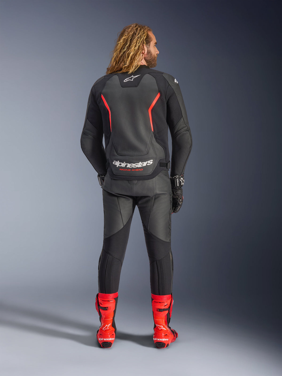 Alpinestars GP Force V2 Leather Jacket, Black and Red Fluo, seen from behind, worn with matching black leather pants and bright red racing boots, featuring red accent lines and bold white Alpinestars branding on the back, professional sport motorcycle riding outfit.