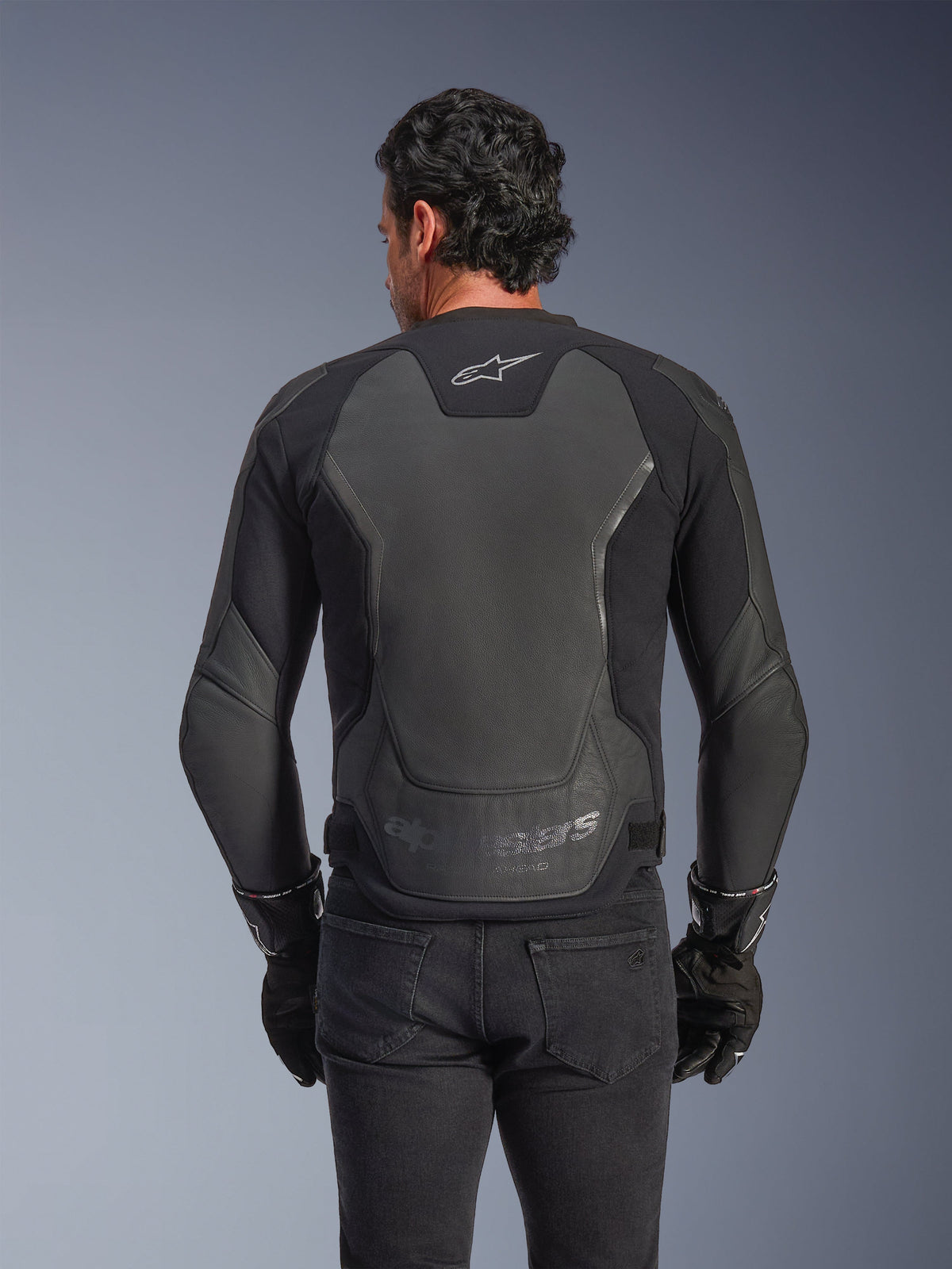 Alpinestars GP Force V2 lederen jas, zwart, achteraanzicht van een man die de jas draagt met zwarte handschoenen en jeans, voorzien van grote lederen beschermingspanelen op de rug, Alpinestars-logo op de bovenrug en reliëfopdruk bij de taille, sportieve motorkleding