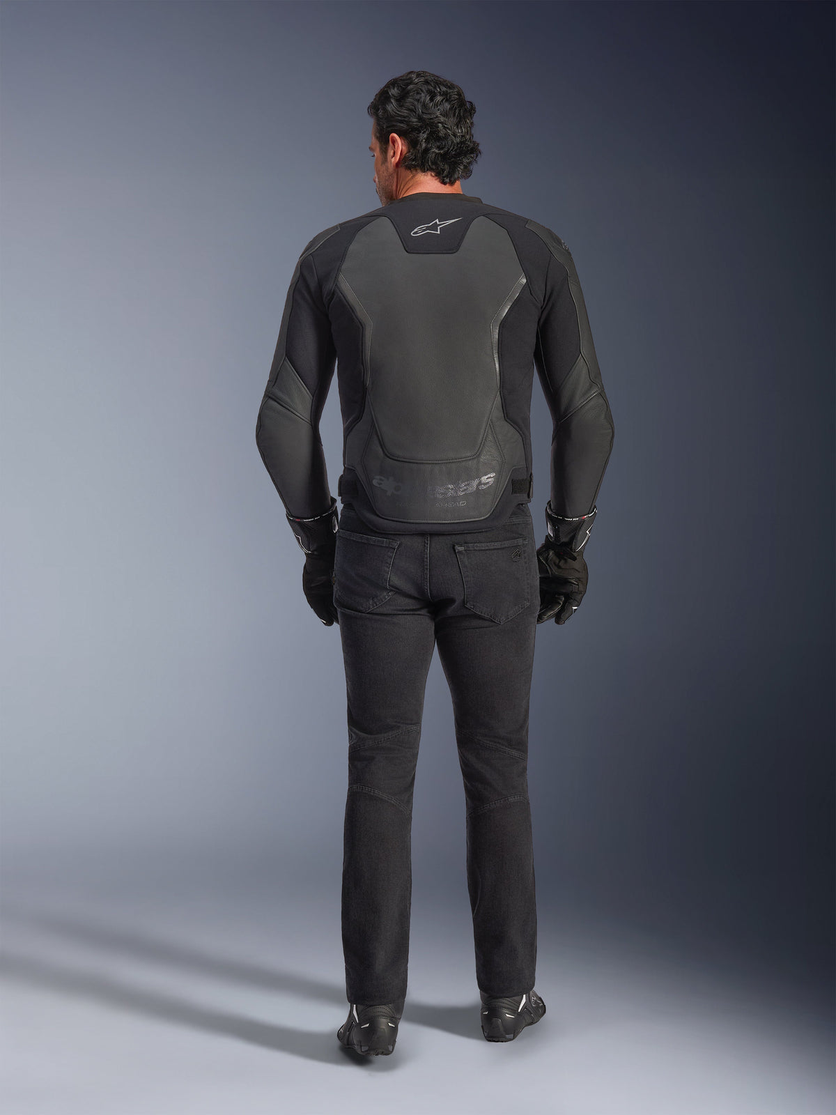 Alpinestars GP Force V2 Leren Jas, zwart, achteraanzicht, premium sportmotorjas met leer-textielconstructie, gedragen met zwarte motorjeans en handschoenen, voorzien van beschermende schouderpanelen en merkdetails
