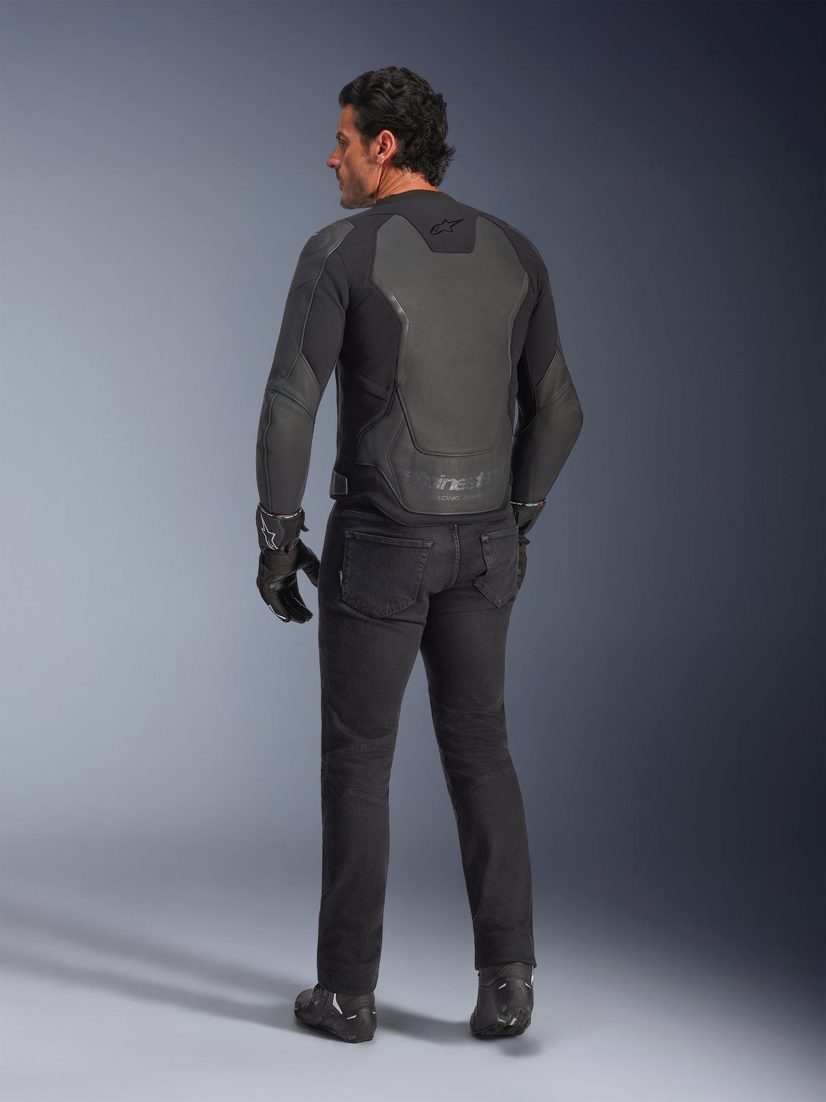 Achteraanzicht van een model die de Alpinestars GP Force V2 leren jas draagt in Black Black, voorzien van een meerpanelige leren constructie en sportieve pasvorm, gecombineerd met een zwarte motorbroek, handschoenen en laarzen voor een complete, op motorracen geïnspireerde look.