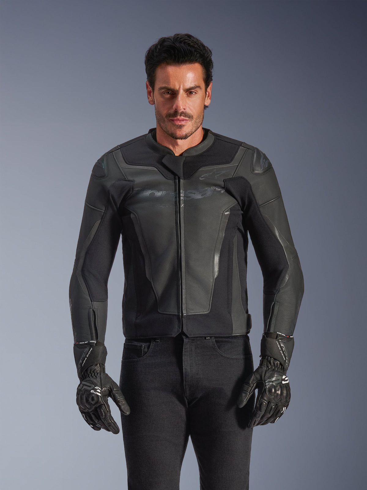 Alpinestars GP Force V2 leren jas, zwart en donkergrijs, motorracejas, man draagt een nauwsluitend leren ontwerp met rits aan de voorzijde, gecombineerd met zwarte getextureerde handschoenen, moderne beschermende uitrusting, hoogwaardige leren constructie