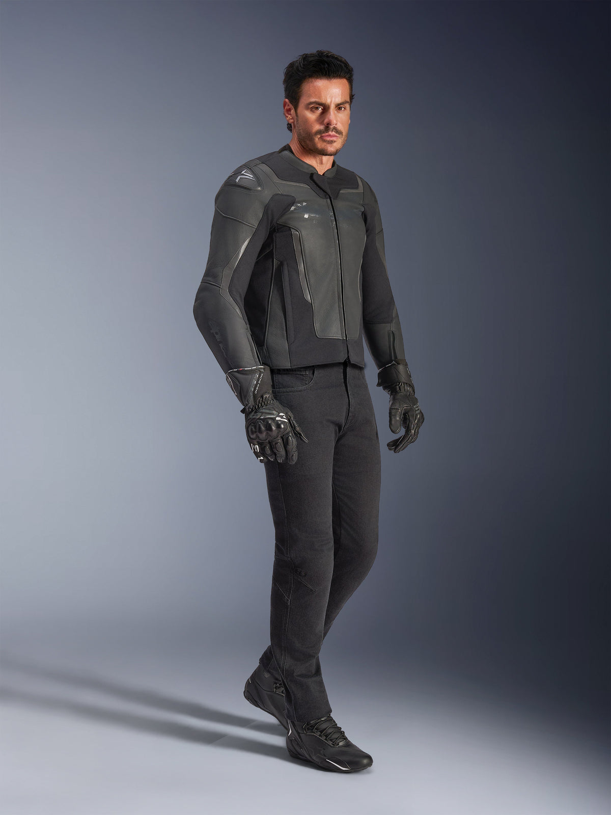 Man die de Alpinestars GP Force V2 leren jas in het zwart draagt, gecombineerd met een zwarte broek en motorhandschoenen, volledige studio-opname, strakke runderleren constructie met geïntegreerde schouder- en elleboogbescherming, professionele sportmotor kleding