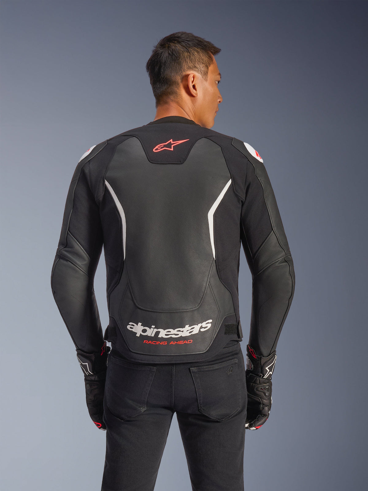 Alpinestars GP Force V2 Leren Jas, achteraanzicht, zwart en wit, voorzien van een rood sterlogo op de bovenrug en witte Alpinestars-branding met 'Racing Ahead'-tekst, gedragen met zwarte broek en bijpassende handschoenen, sportieve leren motorjas