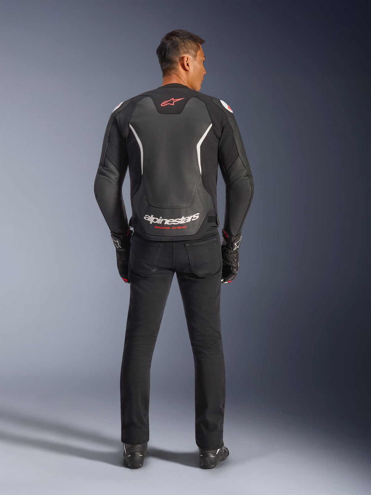 Alpinestars GP Force V2 Leren Jas, zwart en wit, achteraanzicht, gedragen door een rijder met zwarte broek en handschoenen, met groot wit logo op de onderrug en rode accenten, hoogwaardige rundleer constructie voor motorfiets weguitrusting