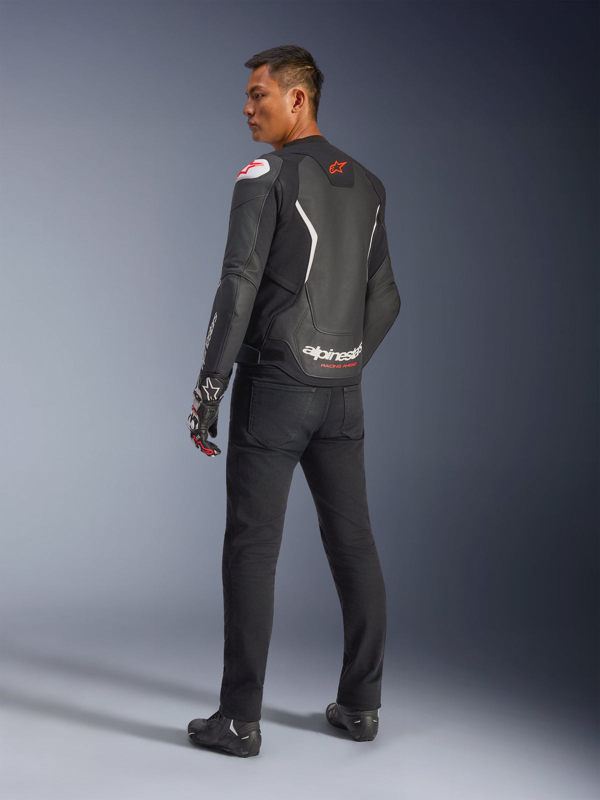 Alpinestars GP Force V2 Leren Jas, zwart en wit, achteraanzicht met leren constructie, witte accenten en rode logo's, gedragen met zwarte broek en racehandschoenen, met opvallende Alpinestars-branding op de onderrug, sportieve motoruitrusting