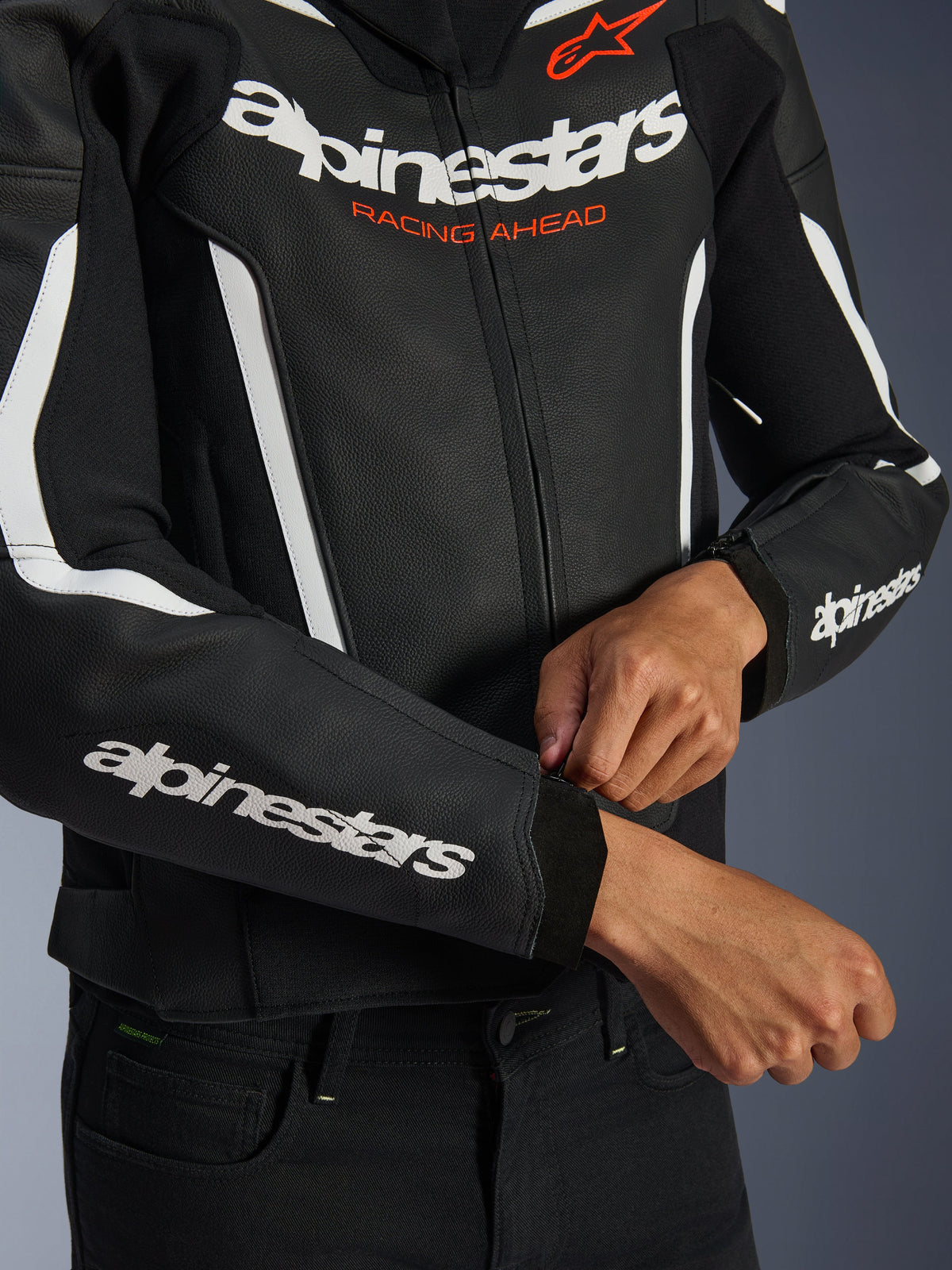 Alpinestars GP Force V2 Leren Jas, zwart en wit, close-up van een rijder die de ritssluiting van de manchet verstelt, witte branding op de borst en mouwen met oranje 'Racing Ahead' tekst, premium rundleer voor sportief motorrijden