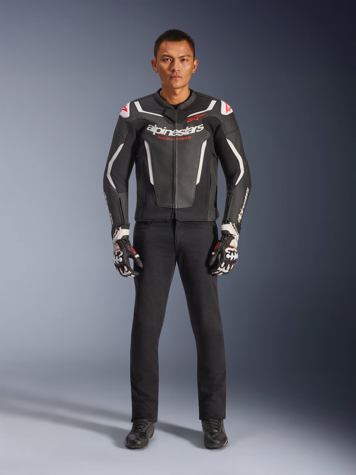 Mannelijk model draagt de Alpinestars GP Force V2 leren jas in zwart en wit, gecombineerd met een zwarte motorbroek en zwart-witte handschoenen, voorzien van opvallende Alpinestars-branding en rode schouderaccenten, professionele sportieve motoruitrusting