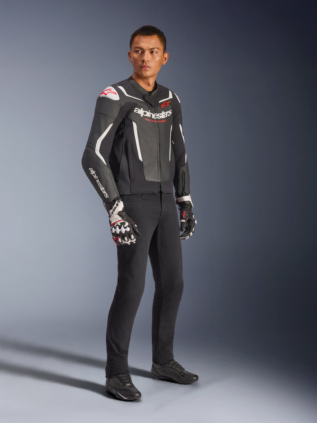Alpinestars GP Force V2 Leren Jas, zwart en wit, gedragen door een mannelijk model met zwarte motorbroek en technische handschoenen, voorzien van rode Alpinestars-logo's, sportieve leren motorjas voor straatgebruik