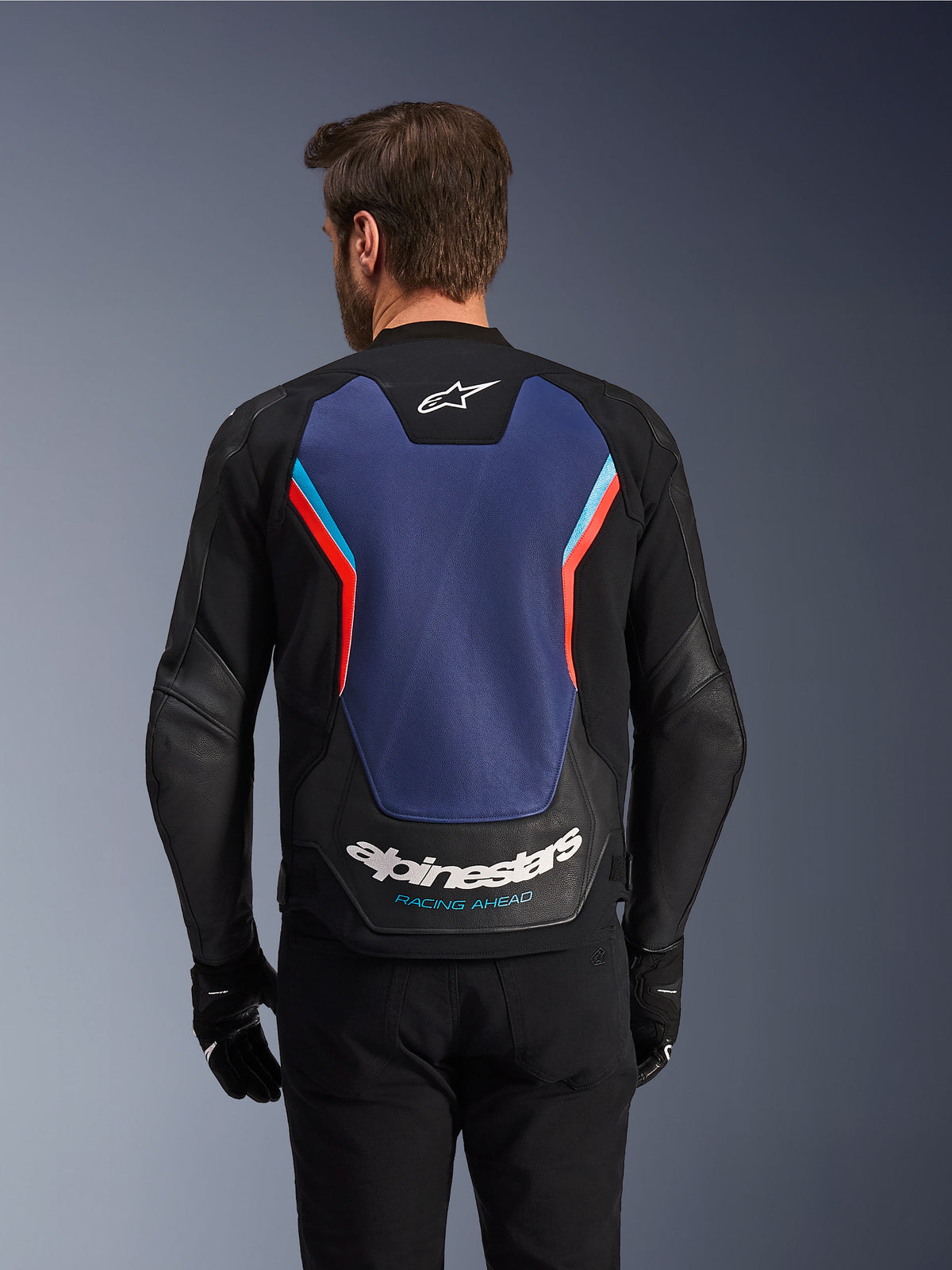 Achteraanzicht van de Alpinestars GP Force V2 leren jas in zwart, blauw en fluo rood, met een groot blauw rugpaneel, fluorescerende rode en lichtblauwe accenten, wit Alpinestars-logo en 'Racing Ahead'-branding op de onderrug, gedragen door een rijder met zwarte handschoenen en broek.