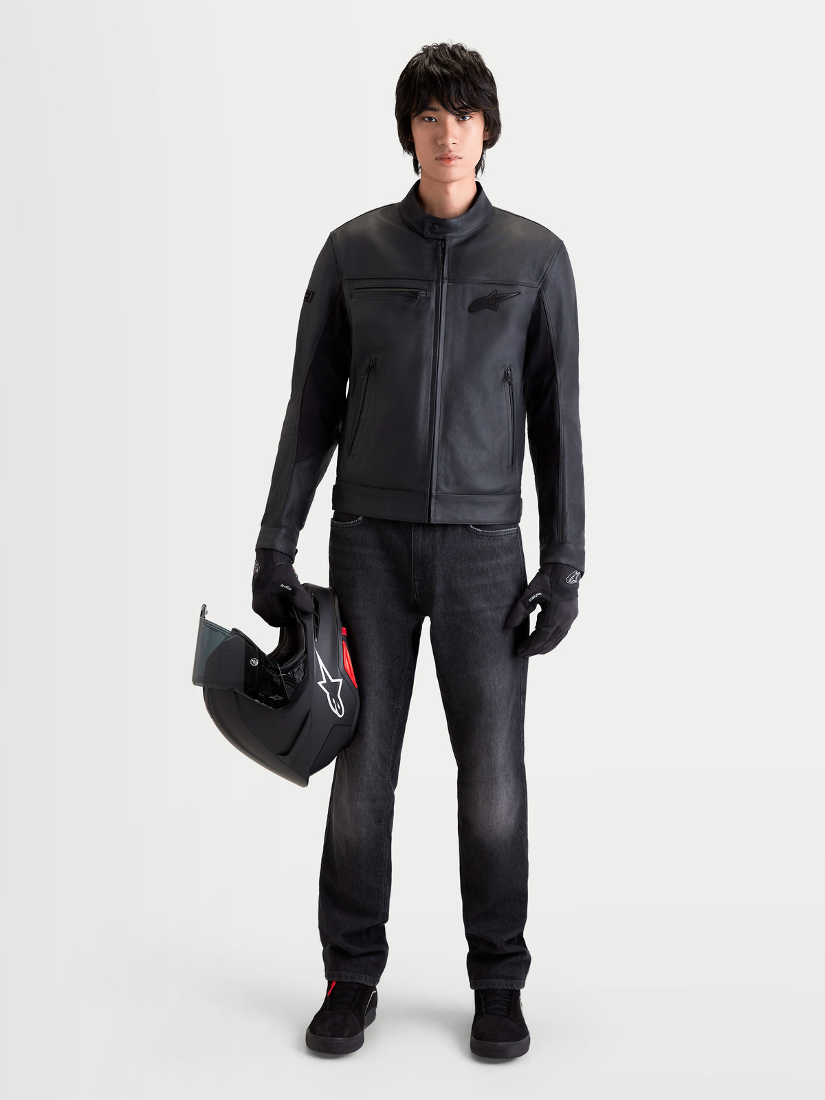 Alpinestars Maver X Leren jas, urban herenmotorjas, zwart, vooraanzicht op model met bijpassende handschoenen en donkere jeans terwijl hij een helm vasthoudt
