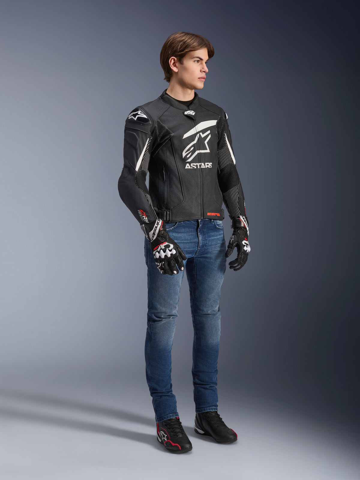 Alpinestars GP Plus R V4 Rideknit Leren Jas, motorleren jas, zwart en wit, gedragen door een model met blauwe jeans en zwarte handschoenen, met een opvallend wit Astars-logo op de borst, geperforeerde panelen en witte accenten op de schouders.