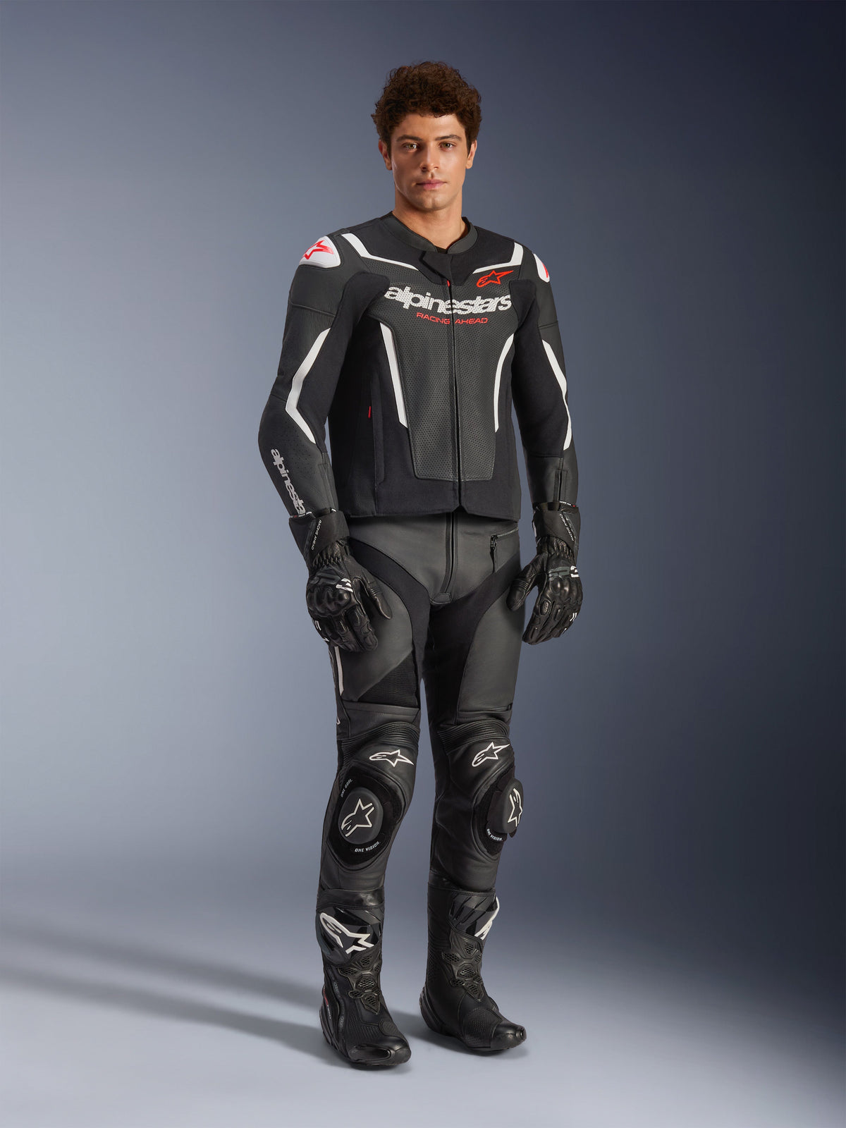 Alpinestars GP Force V2 Air Leather Jacket in Black and White, worn by a male rider with matching black racing pants, gloves, and boots, featuring perforated leather panels, white accents, and red Alpinestars logo on the chest, sport motorcycle riding gear