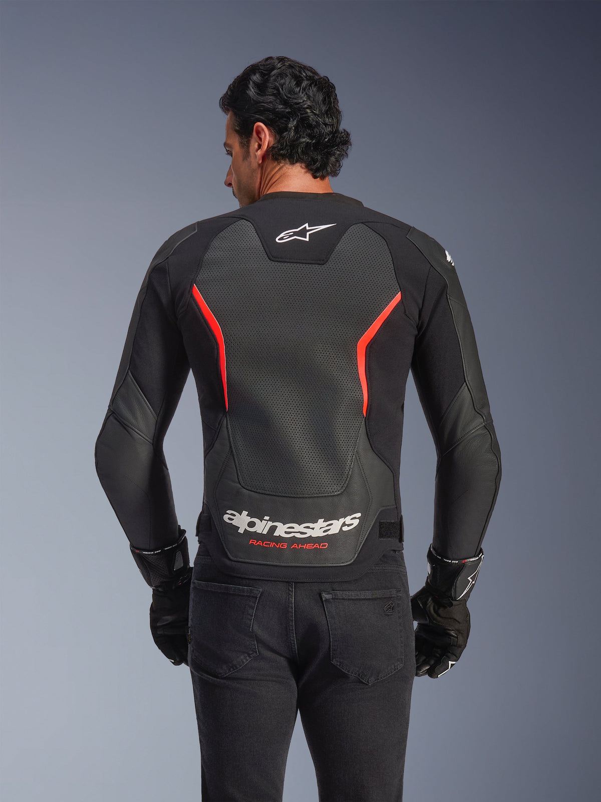 Alpinestars GP Force V2 Air Leather Jacket, back view, Black and Red Fluo, featuring a large perforated leather back panel for ventilation, 'Alpinestars Racing Ahead' branding on the lower back, worn by a rider with black gloves, performance sport motorcycle gear
