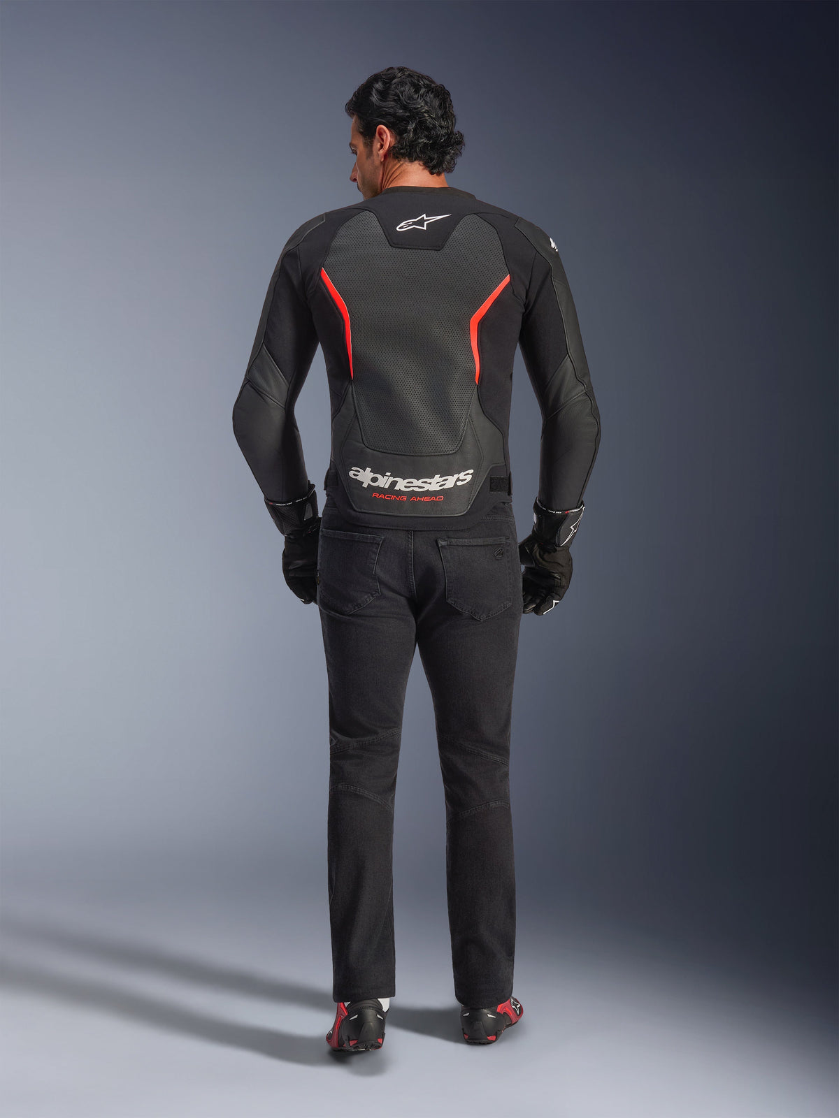 Alpinestars GP Force V2 Air Leather Jacket in Black and Red Fluo, rear view on model, featuring perforated leather back panels, red accents, and Alpinestars branding, styled with black riding jeans and gloves for a sport motorcycle look.