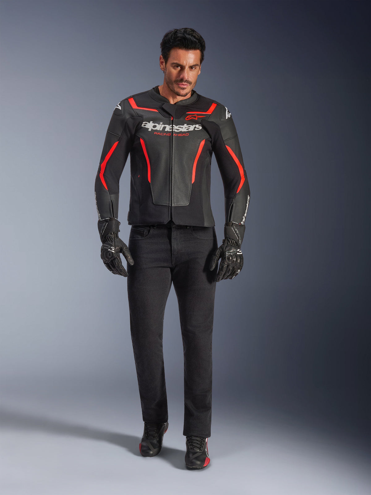 Male rider wearing Alpinestars GP Force V2 Air Leather Jacket, Black and Red Fluo, paired with black riding jeans, gloves, and shoes, featuring perforated leather construction, white logo branding, and sleek red accents for a sport-oriented look