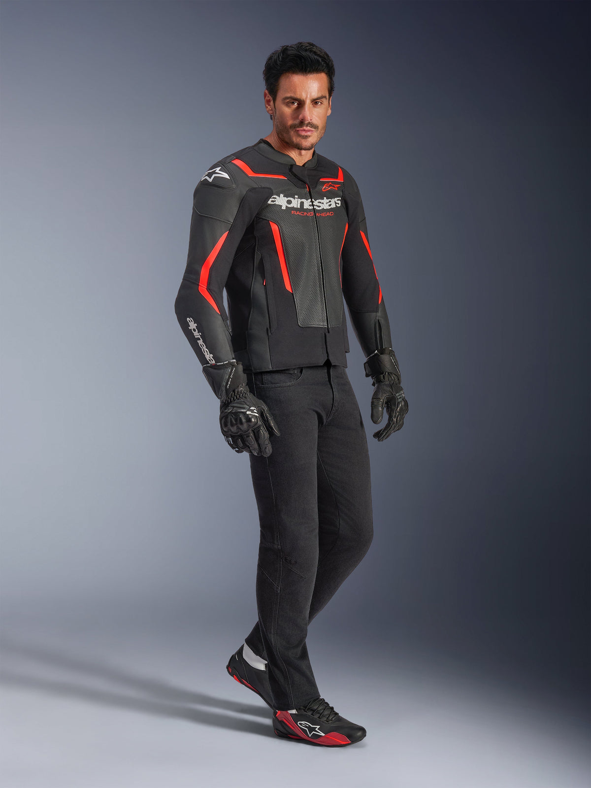 Model wearing Alpinestars GP Force V2 Air Leather Jacket in Black and Red Fluo, paired with black riding pants, gloves, and matching sneakers, featuring perforated leather panels and bold white Alpinestars branding, sport motorcycle gear
