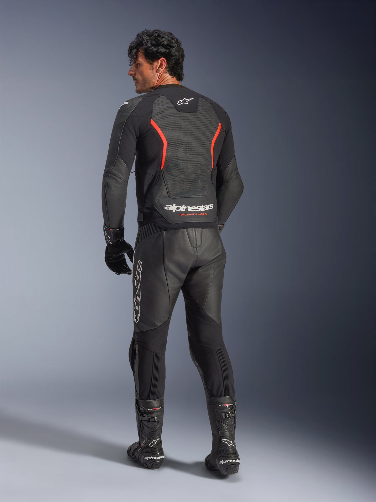 Rear view of a model wearing the Alpinestars GP Force V2 Air Leather Jacket in Black Red Fluo, featuring perforated leather panels and bold Alpinestars branding on the lower back, styled with matching black leather motorcycle pants and boots.