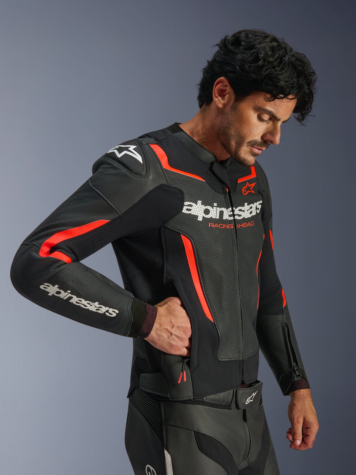 Alpinestars GP Force V2 Air Leather Jacket, Black and Red Fluo, perforated leather motorcycle jacket, model wearing the jacket and reaching into a zippered side pocket, featuring fluorescent red accents and white Alpinestars branding, professional sport riding gear.