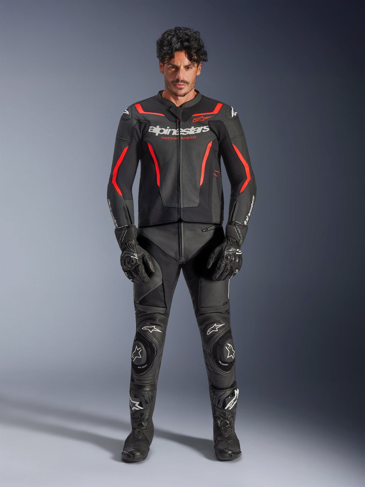Male model wearing Alpinestars GP Force V2 Air Leather Jacket in Black and Red Fluo, featuring perforated chest panels and white branding, paired with black motorcycle leather pants, black gloves, and black boots for a complete racing look.